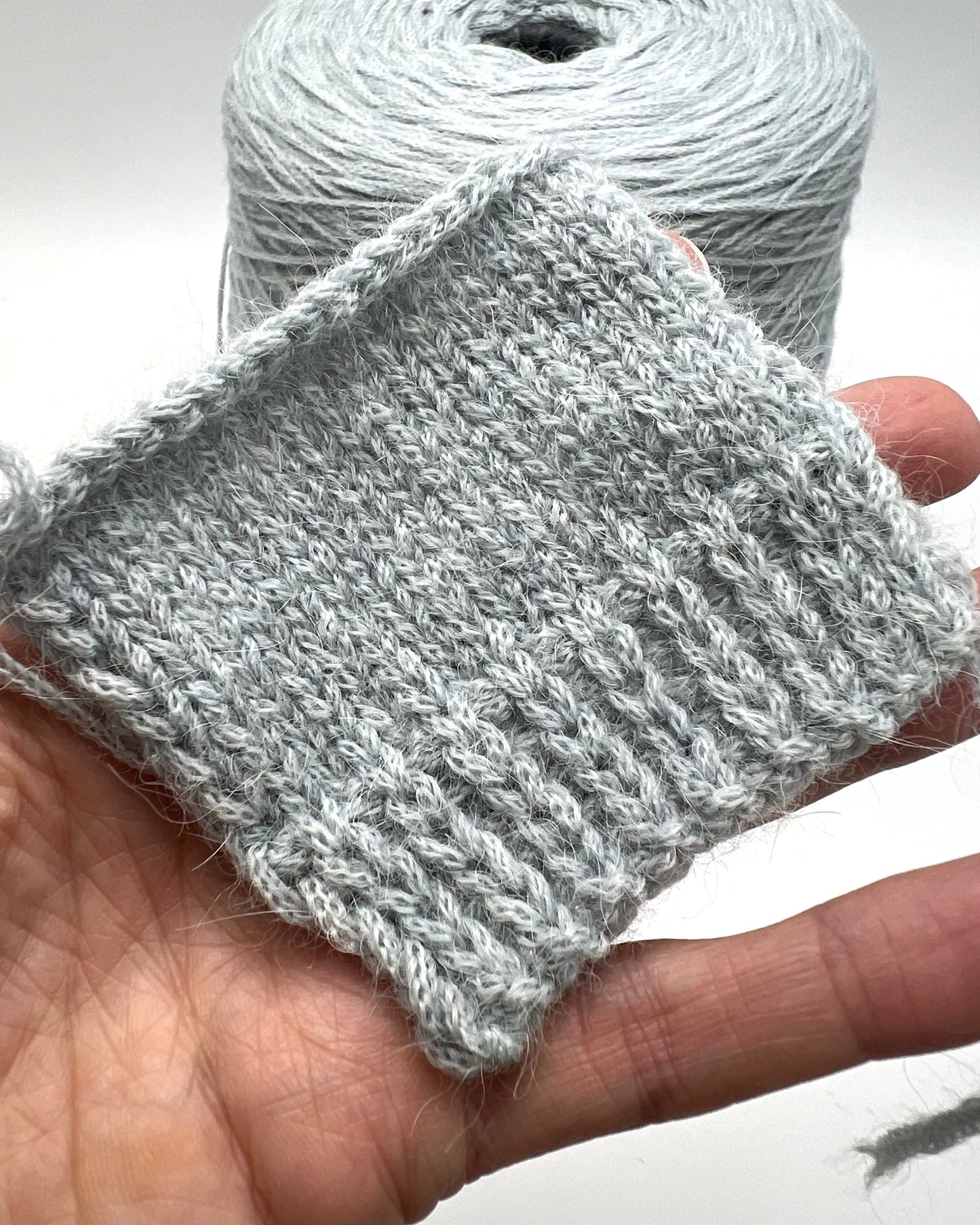 Knitted gray fabric swatch held in a hand with a ball of yarn in the background.