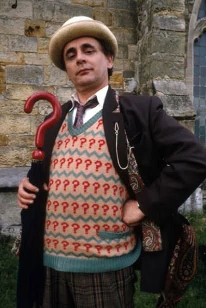 Person wearing a patterned sweater and hat with a red cane, standing in front of a stone wall.