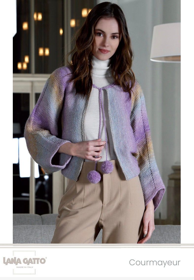 Woman wearing a colorful knitted cardigan with pom-poms indoors, brand 'Lana Grossa' visible.
