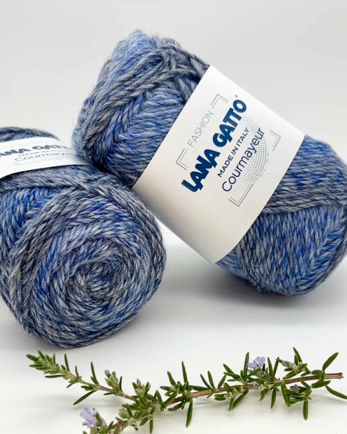 Two balls of blue yarn with a label on a white background, accompanied by a sprig of rosemary.