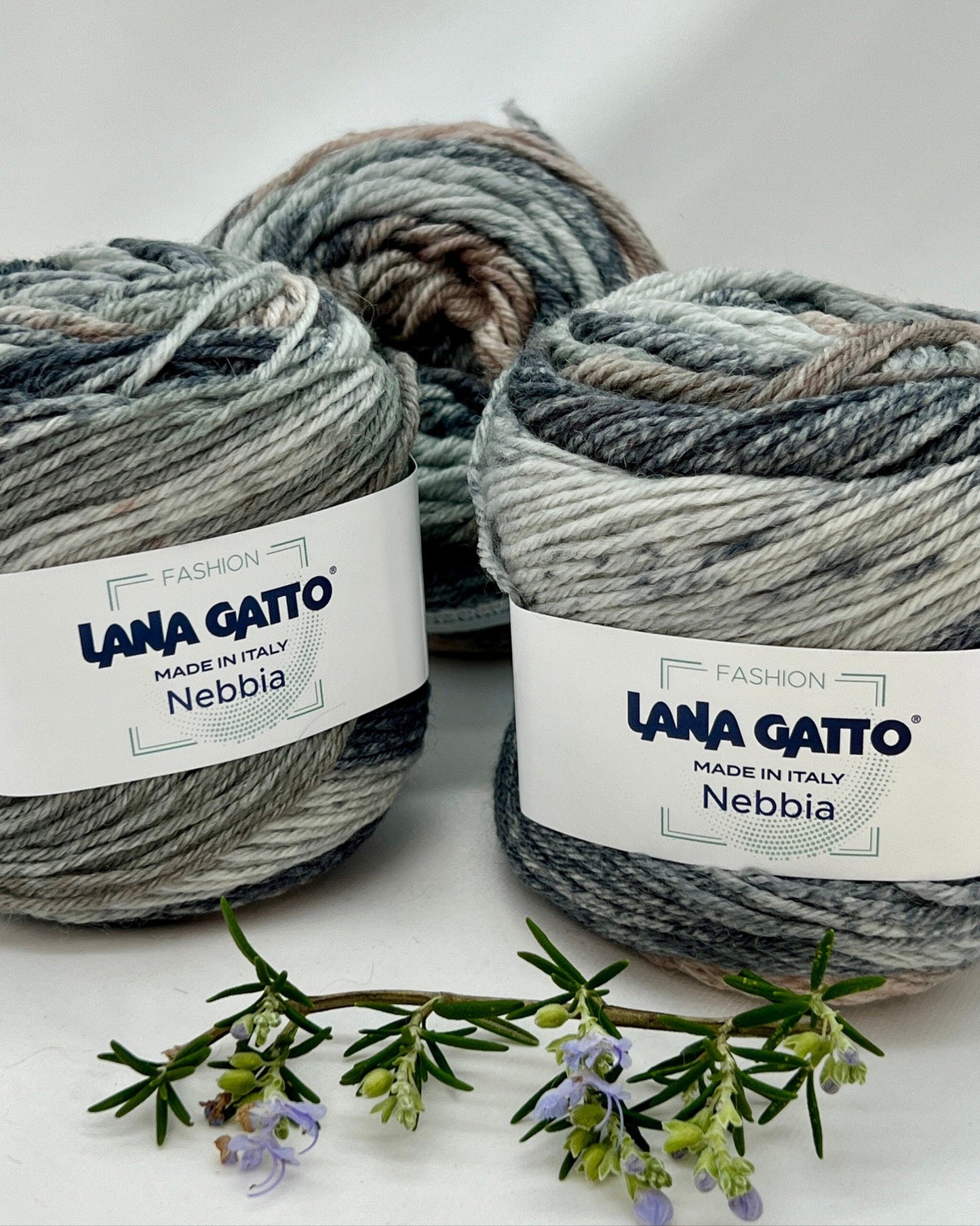 Three balls of Lana Gatto Nebbia yarn on a white background with a sprig of lavender.