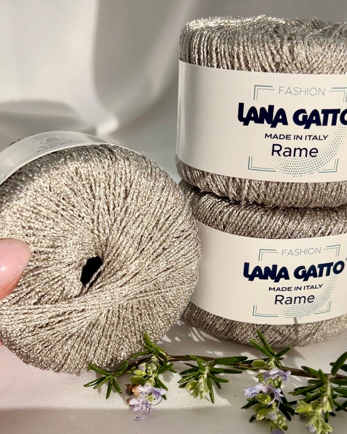 Two balls of Lana Gatto yarn labeled 'Rame' on a light background with a hand and flowers.