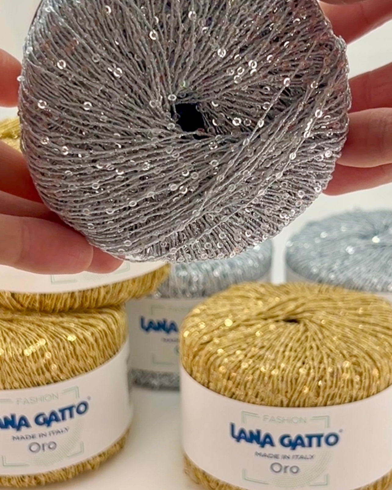 Hand holding a ball of silver yarn with other balls of yarn labeled 'Lana Gatto' in the background.