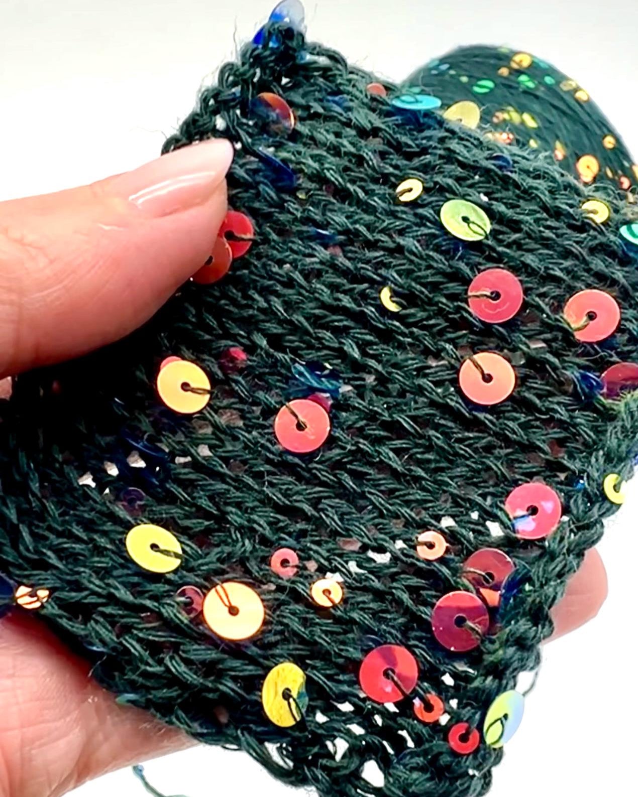Hand holding a green knitted item with colorful sequins on a white background