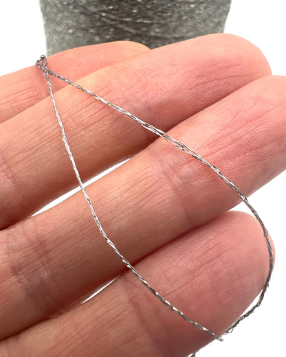 A professional cone of Kyototex MANNA-GROSSA metallic lamé yarn in Brushed Gold. The image shows the yarn's intense shimmer and fine texture, with detail shots of the fiber being held to demonstrate its lace-weight scale. Available at iDOKNiT.com.