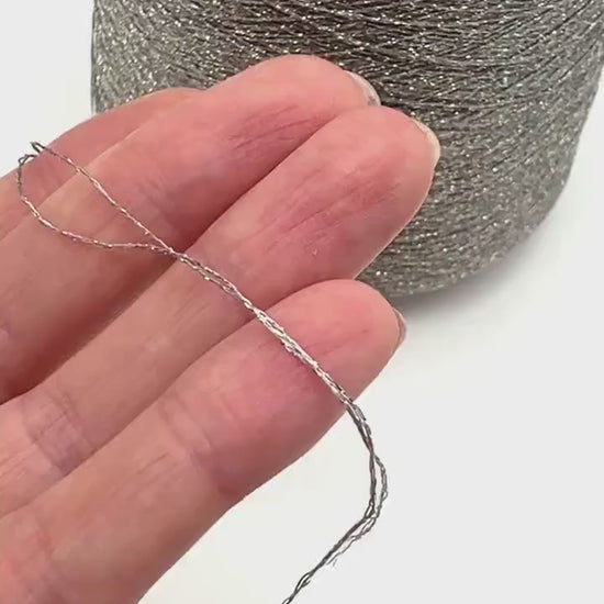 A professional cone of Kyototex MANNA-GROSSA metallic lamé yarn in Brushed Gold. The image shows the yarn's intense shimmer and fine texture, with detail shots of the fiber being held to demonstrate its lace-weight scale. Available at iDOKNiT.com.