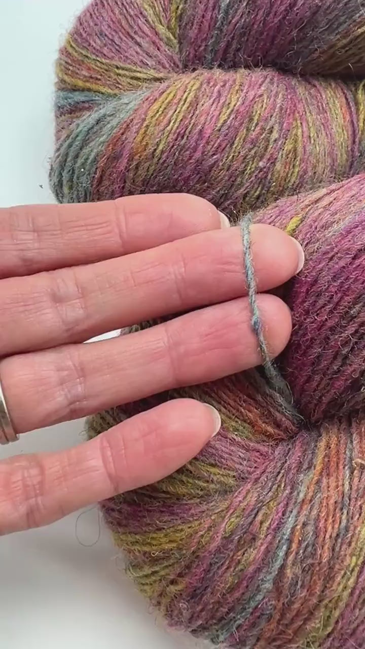 Small-batch dyed heritage skeins of 100% Latvian Dark-Headed Sheep Wool in earthy multicolor gradients like Green-Brown #052. The image shows the rustic single-ply texture and the sheep breed origin, representing the authentic wool traditions of Paces Vilnas Fabrika, Latvia. Available at iDOKNiT.com.