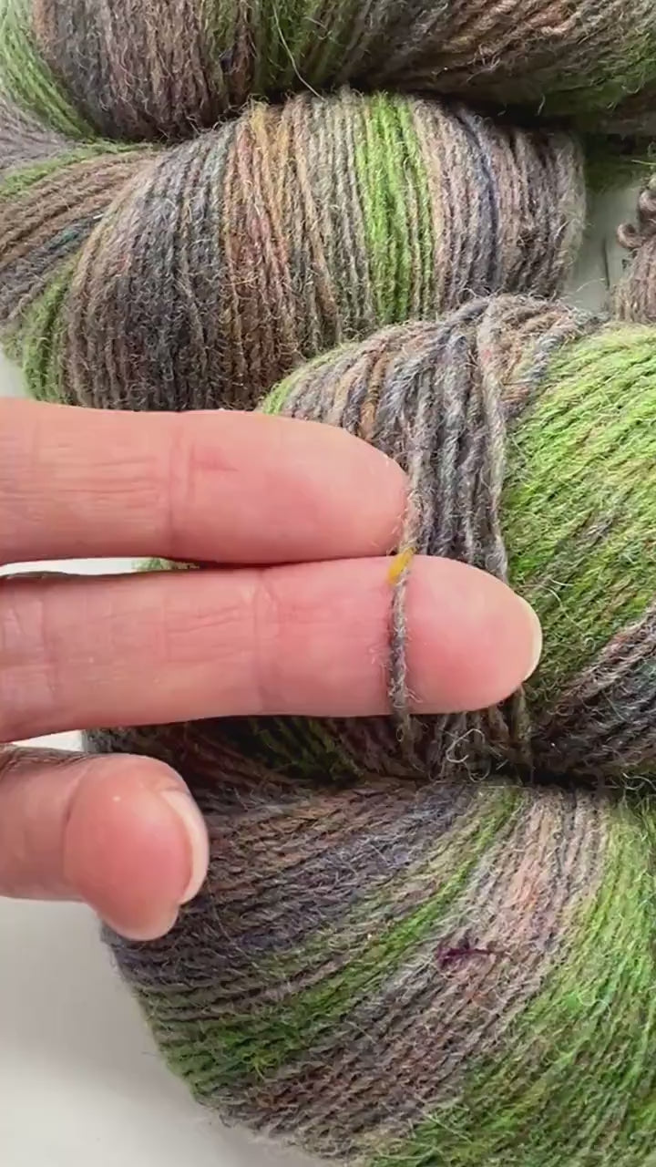 Small-batch dyed heritage skeins of 100% Latvian Dark-Headed Sheep Wool in earthy multicolor gradients like Green-Brown #052. The image shows the rustic single-ply texture and the sheep breed origin, representing the authentic wool traditions of Paces Vilnas Fabrika, Latvia. Available at iDOKNiT.com.